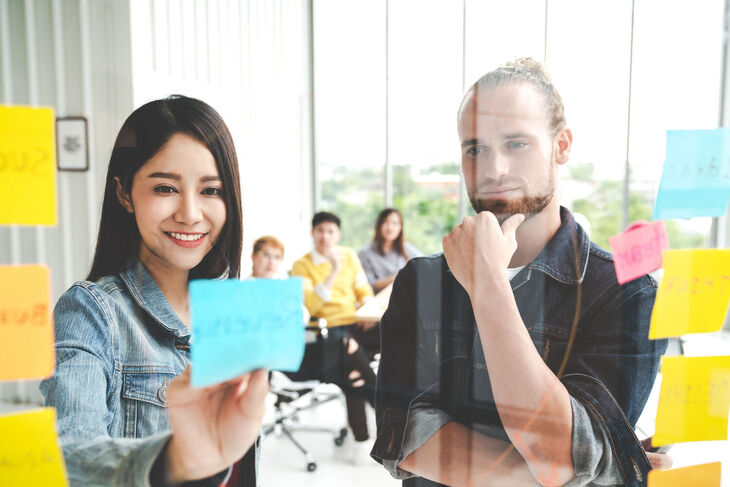 Gruppe von jungen erfolgreichen und kreativen Mitarbeitende lächeln im Team und machen ein Brainstorming für das Modulbaukasten Projekt für Unternehmen. Die Frau klebt ein PostIt mit einer Idee an die Wand, welche begutachtet wird.